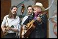 A person playing guitar and singing with a person in a cowboy hat

Description automatically generated